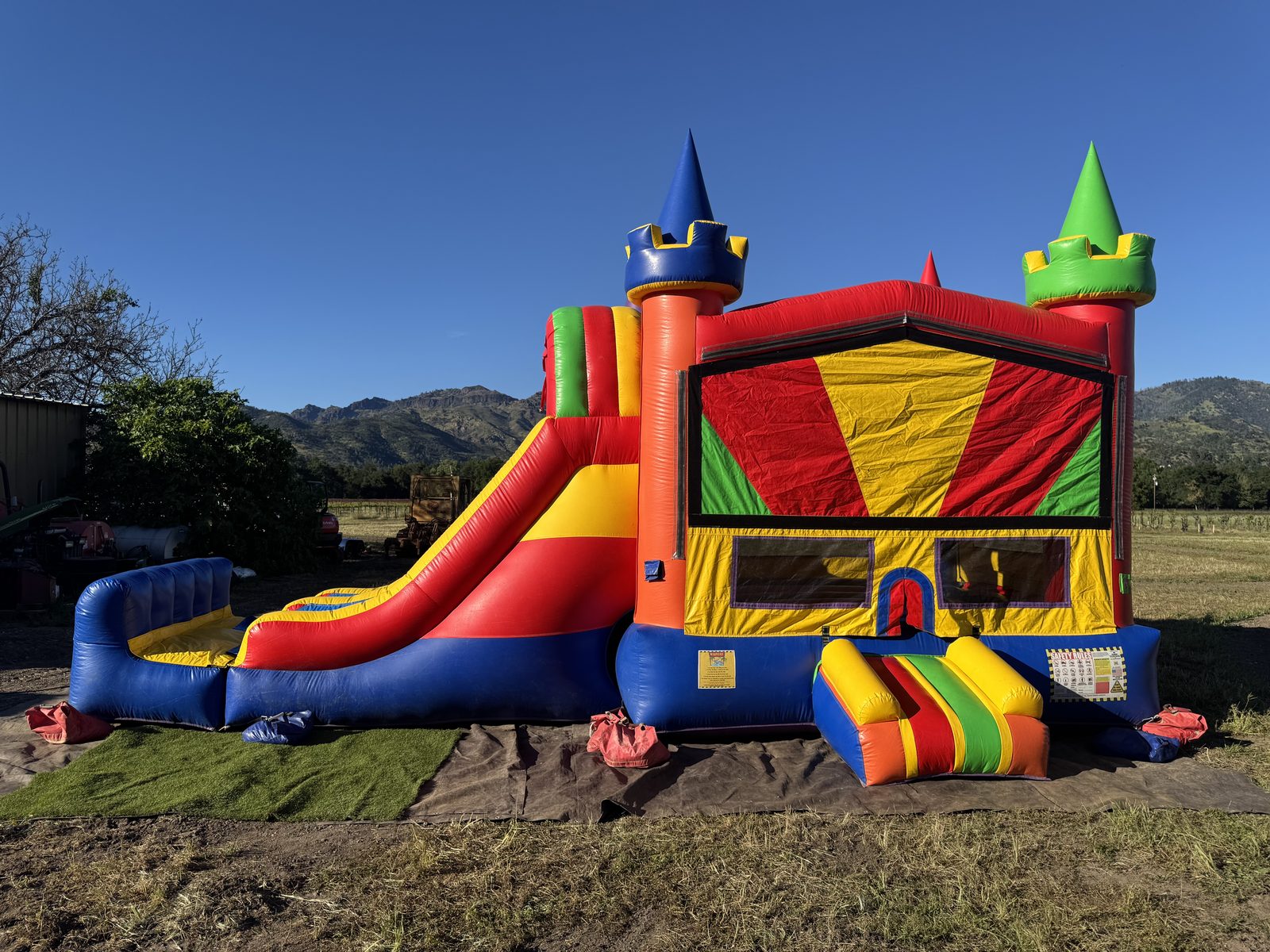 A Toads Jump castle bounce house combo with a slide set up on turf and a tarp in Calistoga, CA, with mountains and a blue sky behind